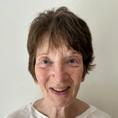 An older woman with short brown hair and blue eyes smiles at the camera, wearing a white shirt, standing in front of a plain, light-colored background.