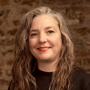 A woman with long wavy gray hair, wearing a black shirt, smiles in front of a rustic brick wall background.