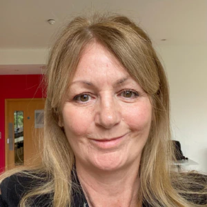 A woman with shoulder-length blonde hair smiles at the camera indoors, wearing a black blazer and necklace. A red wall and a wooden door are visible in the background.