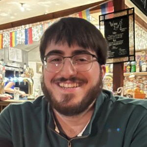 A man with glasses and a beard smiles while sitting at a wooden table in a cozy bar, arms folded. The bar is decorated with colorful flags and a chalkboard menu hangs in the background.