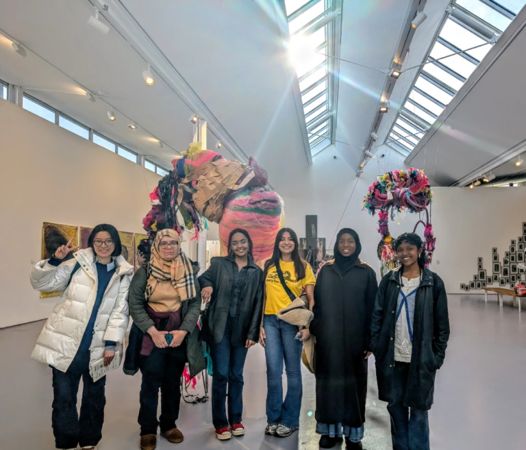 Six women stand smiling in an art gallery with bright lighting, colorful hanging sculptures, and various artworks displayed on white walls in the background.