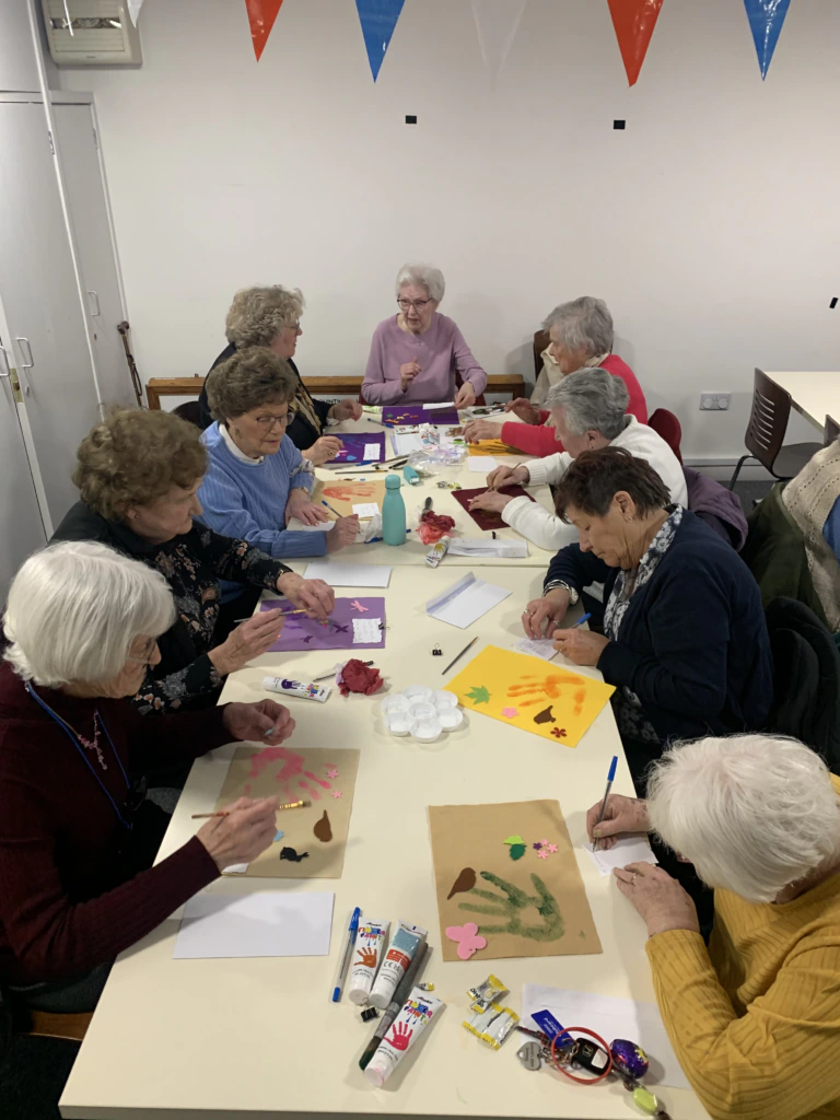 A group of elderly women sit around a table in a craft room, making art with colored paper, brushes, and paint. They appear focused and engaged, with art supplies and finished pieces scattered on the table.