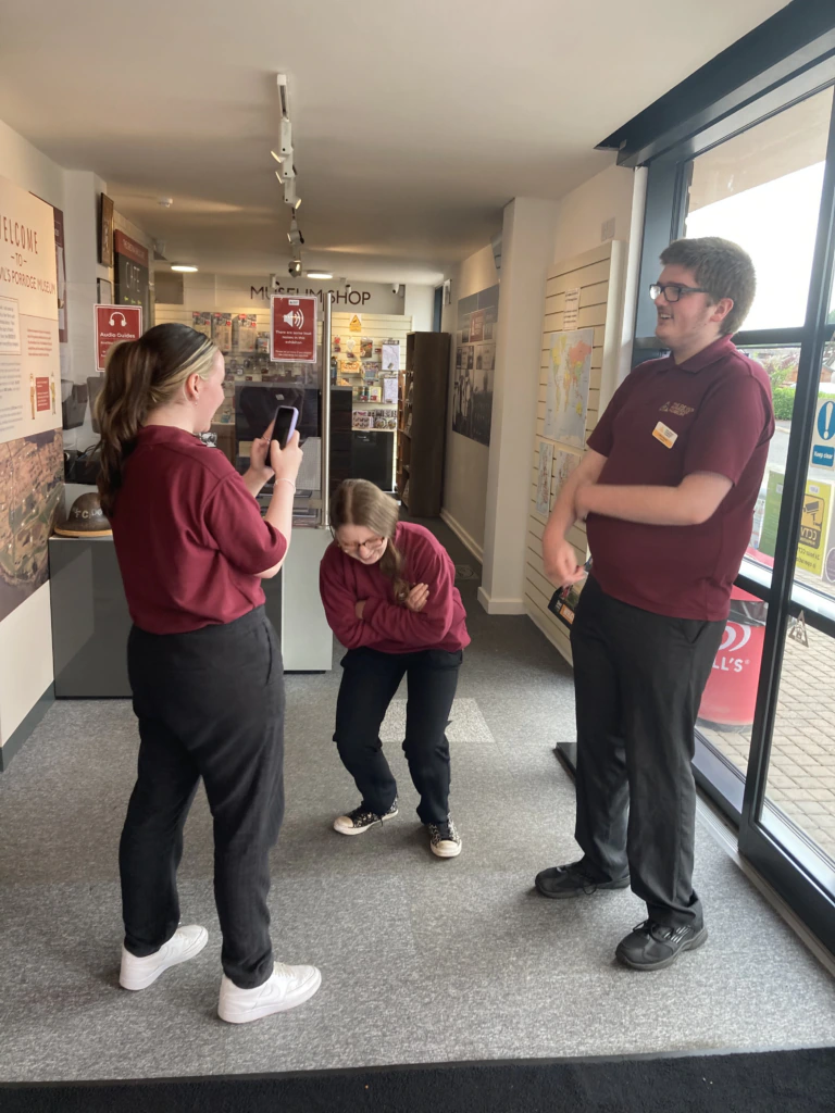 Three people in matching maroon shirts pose indoors; one stands and films with a phone, another laughs and crouches, while the third stands smiling with arms crossed near large windows.