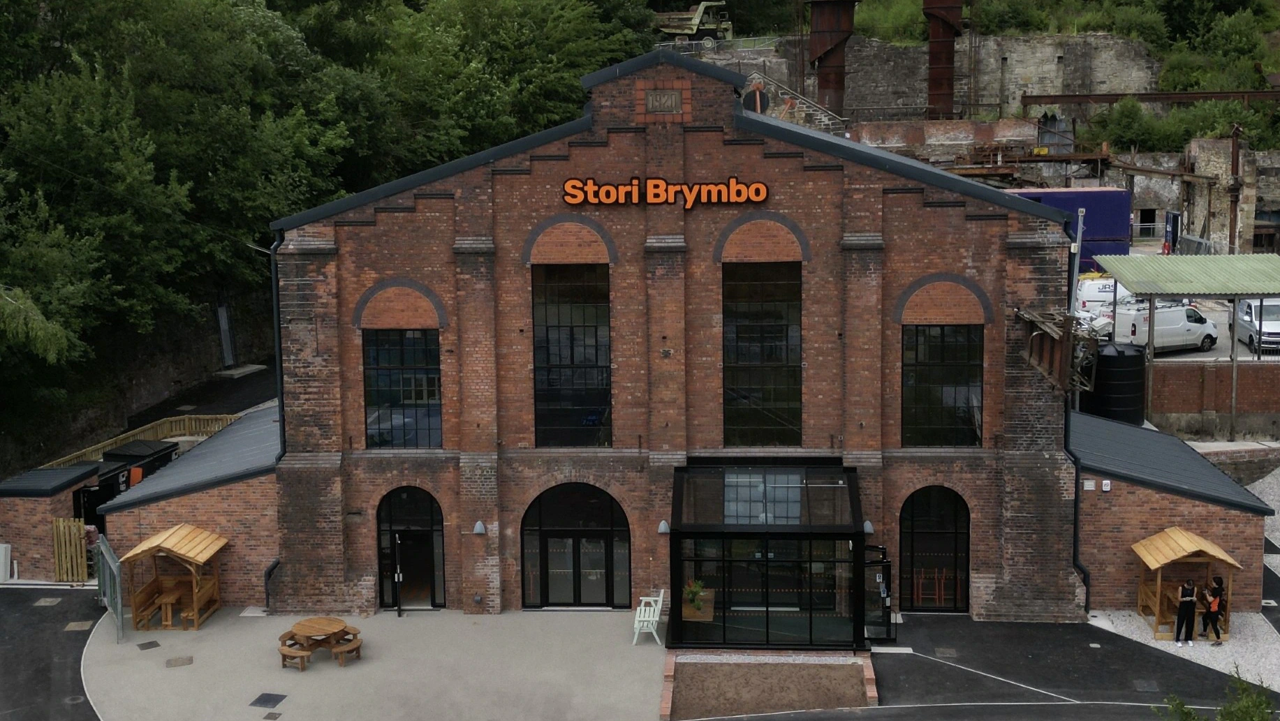 A large brick industrial building labeled Stori Brymbo stands among green trees, with outdoor seating and people wearing orange vests near the entrance.