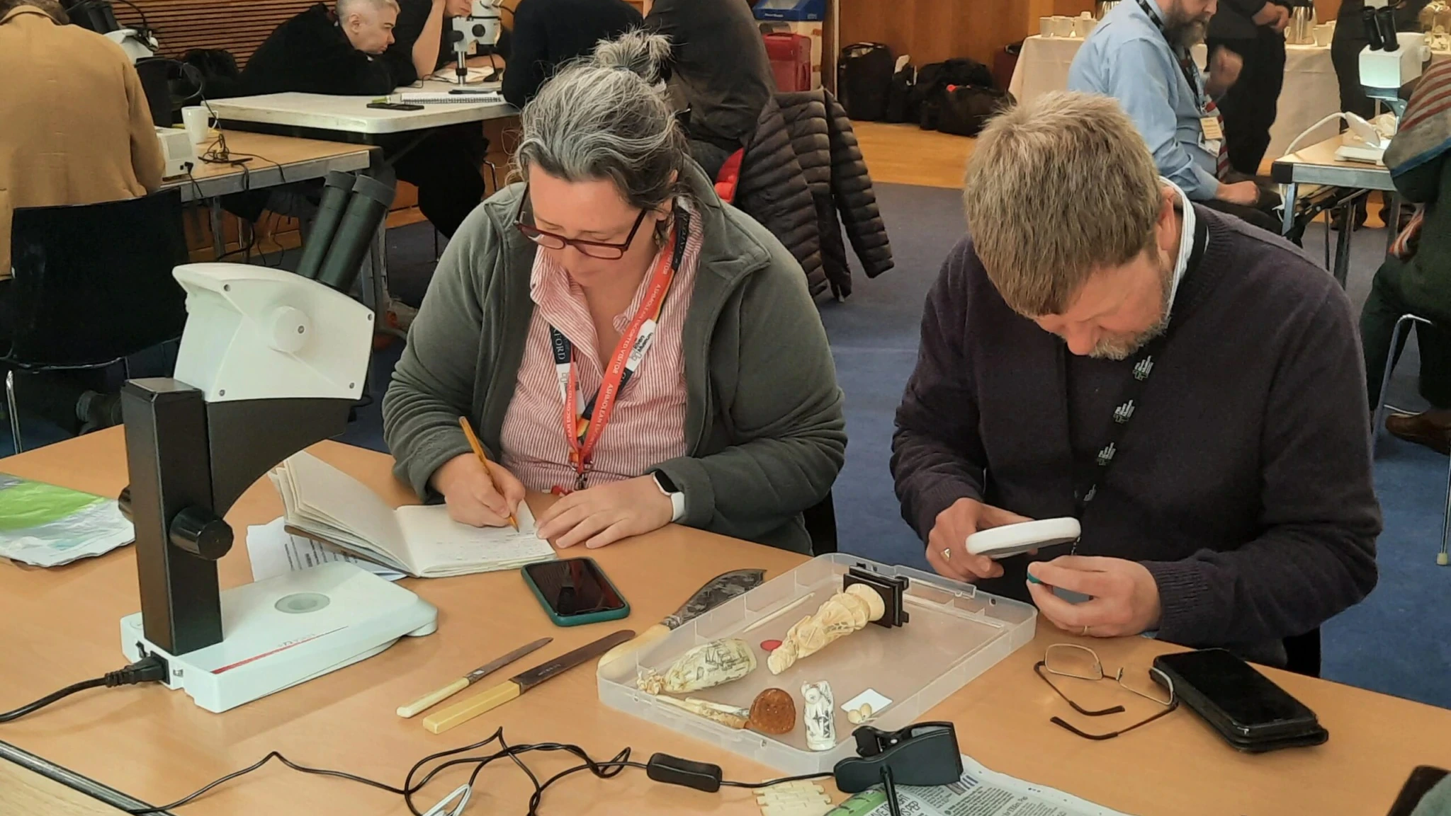 Two people sit at a table examining artifacts and taking notes. Scientific equipment, papers, and tools are spread out, while others work at tables in the background in a conference or workshop setting.
