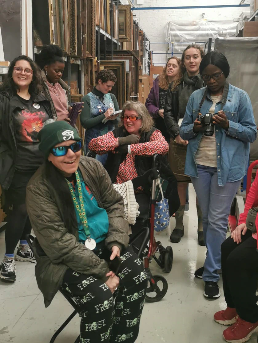A group of people poses and smiles inside an art storage room with shelves of framed artwork. One person sits on a chair in front, wearing sunglasses and a hat; others stand behind, some holding cameras.
