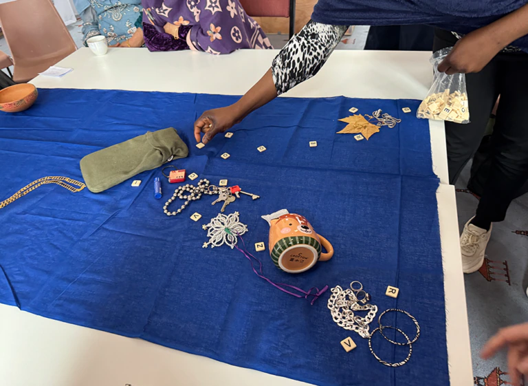 People arrange jewelry, a mug, letter tiles, and small objects on a blue cloth on a table. One person holds a bag of tiles, while others interact with the items. A bowl and fabric pouch are nearby.