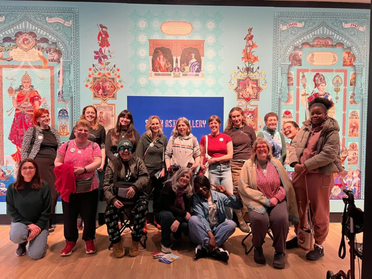A group of people pose and smile together indoors in front of a colorful, ornate wall display and a blue sign. Some are standing, some kneeling, and one person has a walking aid.
