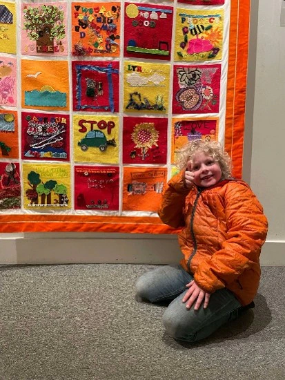 A child in an orange jacket kneels and smiles beside a colorful quilt, which is made up of many squares, each decorated with different images and designs.