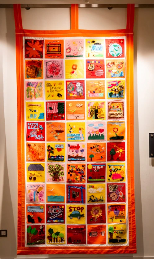A colorful quilt with 42 squares, each decorated with unique drawings and crafts. The quilt has a bright orange border and hangs on a wall.