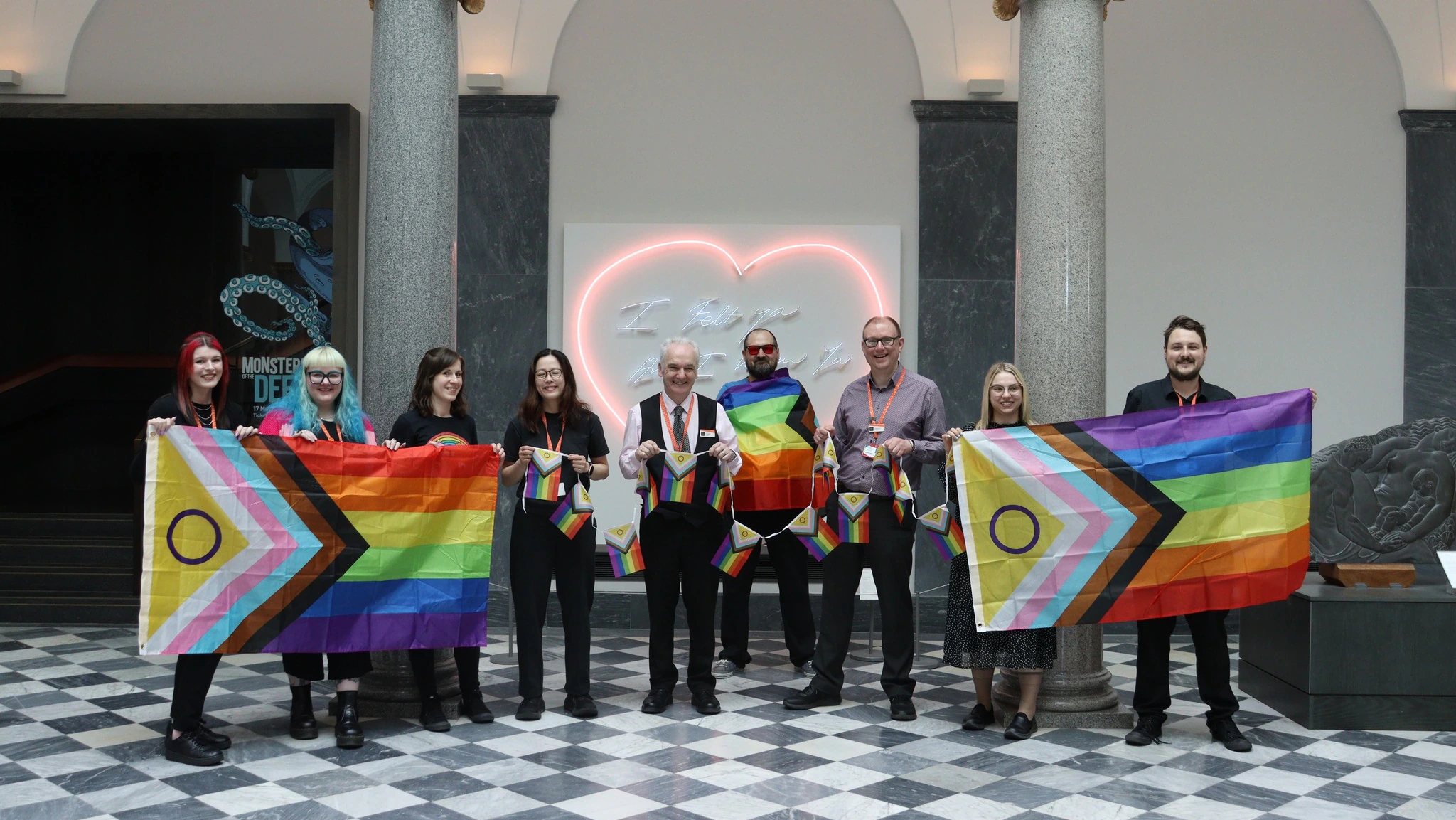 Eight people stand indoors holding Progress Pride flags and bunting. Behind them are marble columns, grey walls, and a neon heart sign. The floor features a black and white checkerboard pattern.