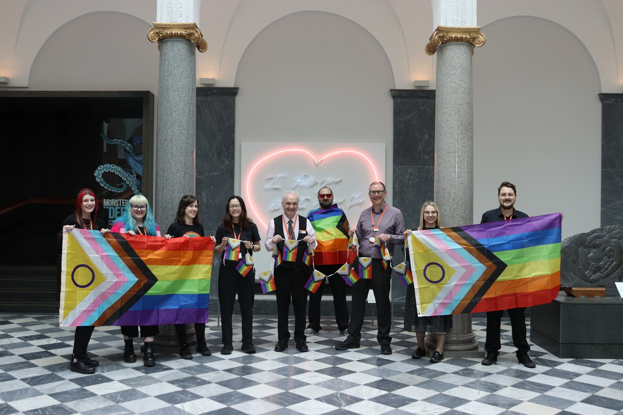 Eight people stand indoors holding Progress Pride flags and bunting. Behind them are marble columns, grey walls, and a neon heart sign. The floor features a black and white checkerboard pattern.