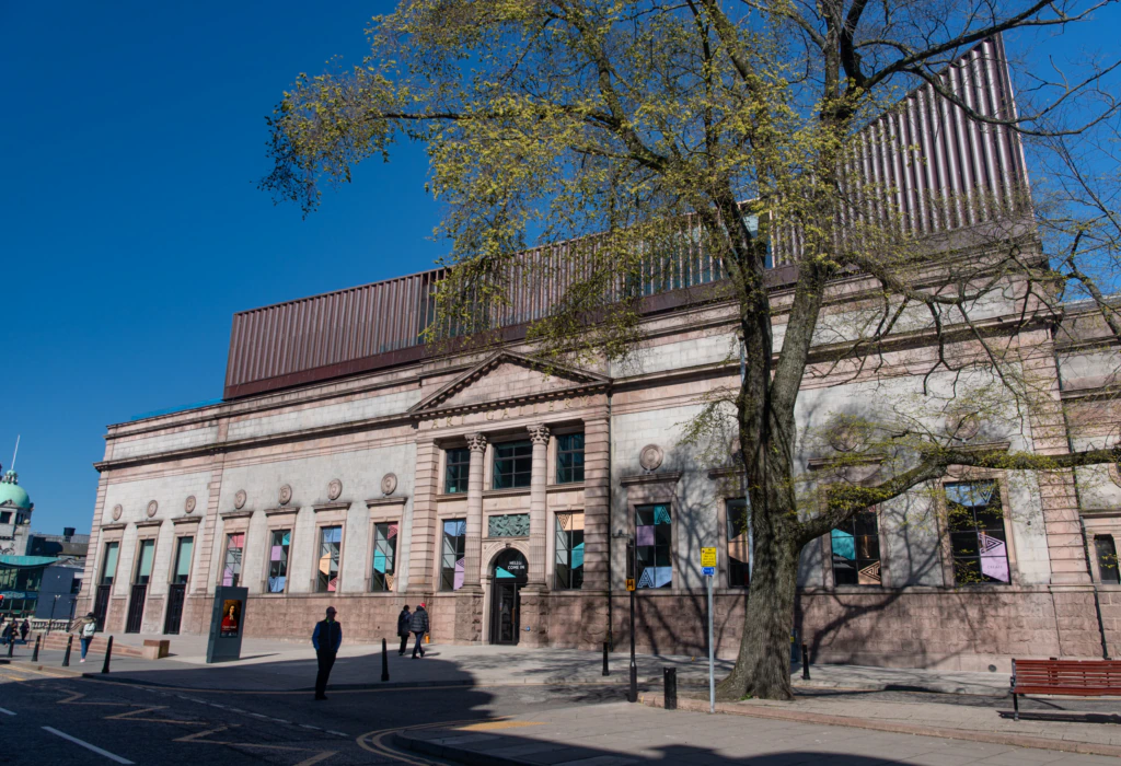 A large stone building with columns and tall windows stands under a clear blue sky. A tree with new leaves partially shades the sidewalk, while a few people walk nearby.