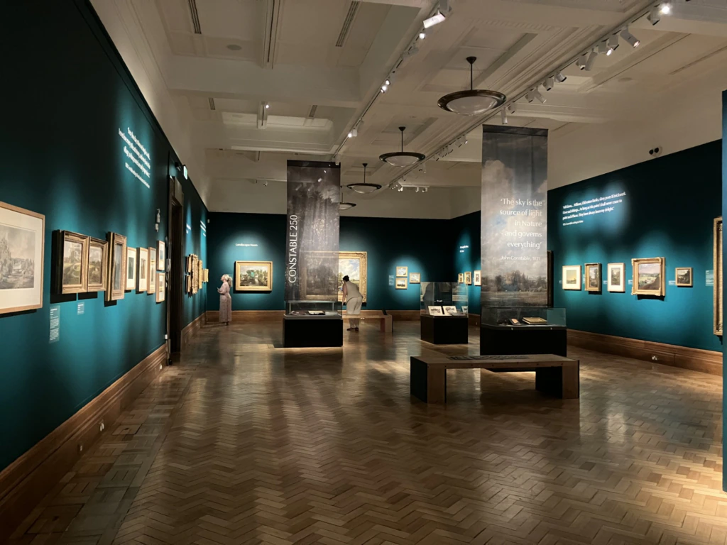 Art gallery with green-blue walls, framed paintings, and display cases. Two people observe art. Overhead lights illuminate the exhibits. Large banners with text hang from the ceiling. The space has a quiet, contemplative atmosphere.