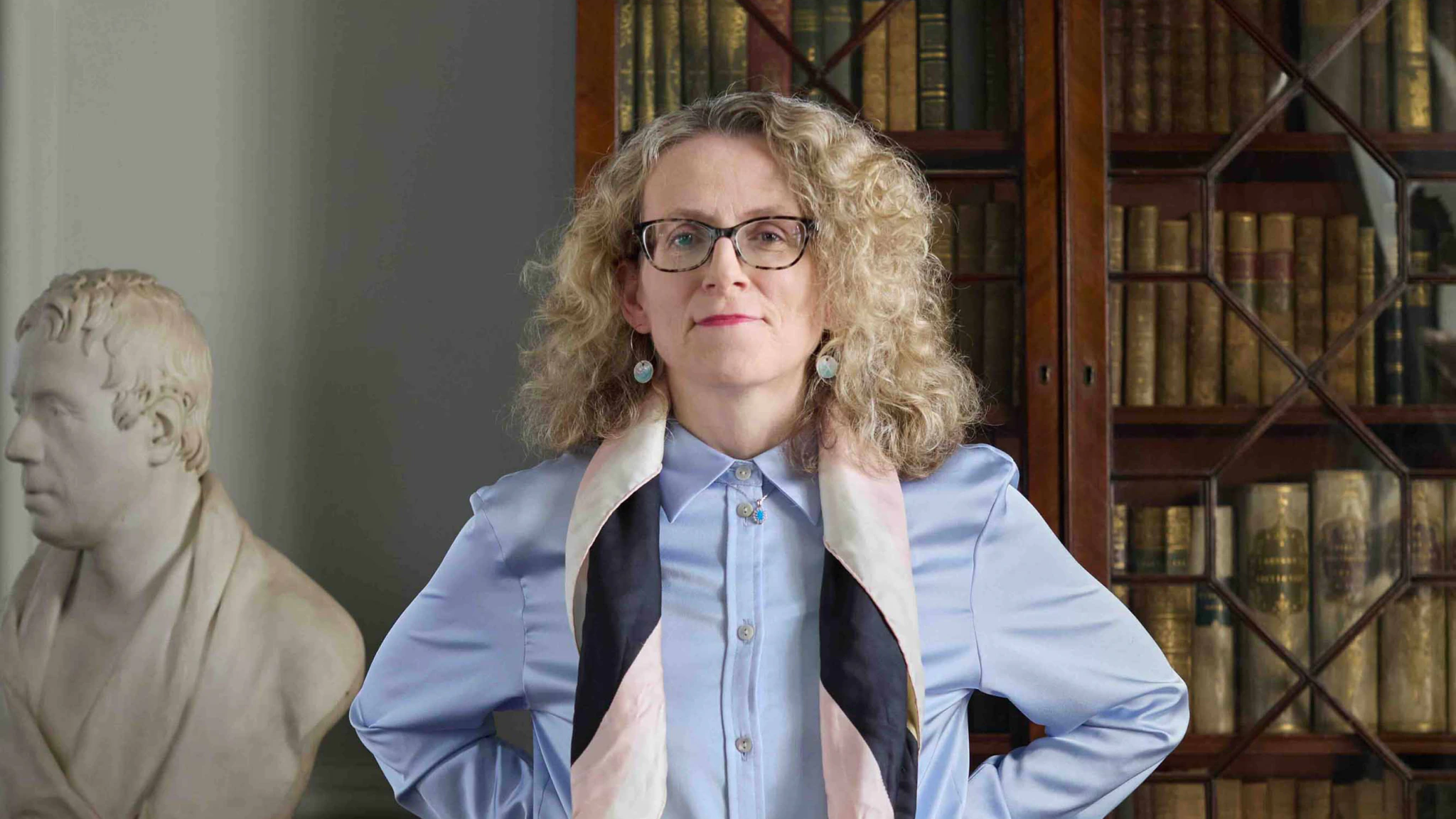 A woman with curly blonde hair and glasses stands confidently in front of bookshelves, wearing a light blue shirt and a patterned scarf. A white bust statue is visible to her left.