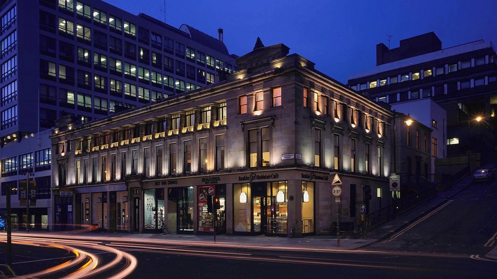 A well-lit building on a city street corner at dusk, with light trails from passing cars and modern office buildings in the background.