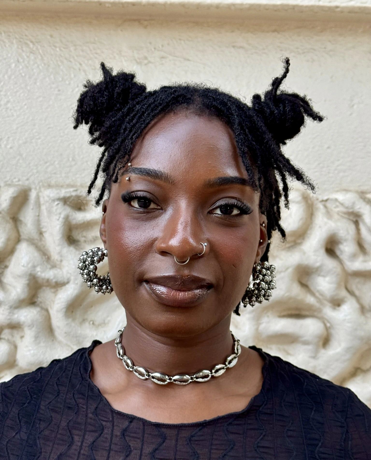 A person with short twisted hair in two buns, hoop earrings, a nose ring, and a shell necklace stands in front of a textured, off-white wall, wearing a black top and looking directly at the camera.