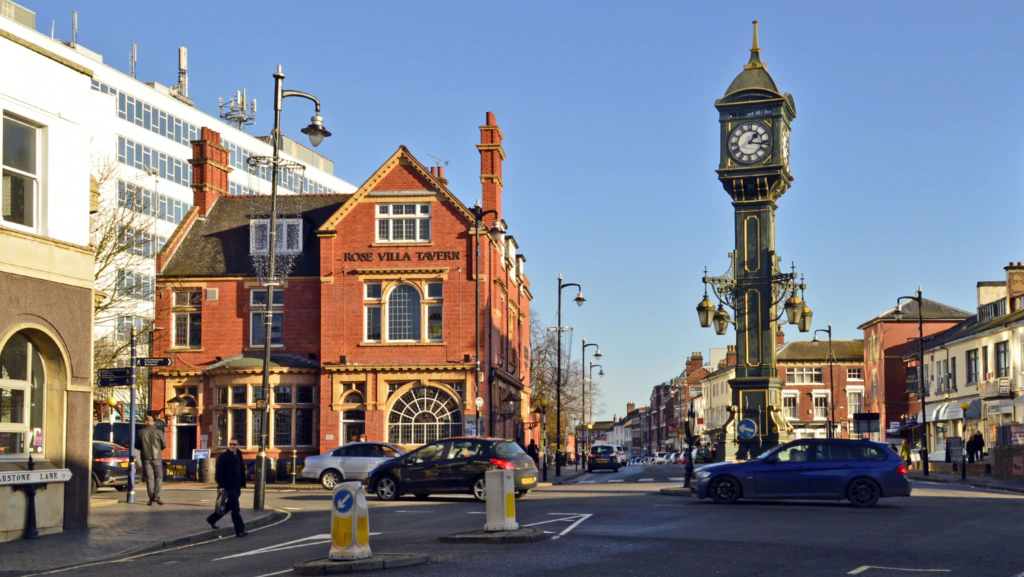 A street scene with the Rose Villa Tavern, a red-brick building, on the left and a tall clock tower at a roundabout. People walk, and cars drive on a sunny day in an urban area.