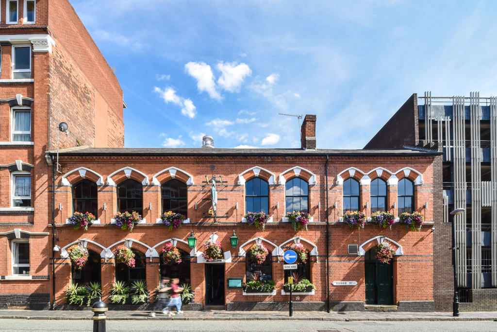 A red-brick building with arched windows, hanging flower baskets, and green lamps. Two people walk past on the sidewalk. Blue sky with clouds above.