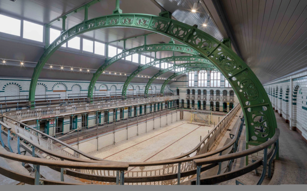 An empty, historic indoor swimming pool with ornate green iron arches, tiled walls, and large windows filling the spacious hall with natural light.