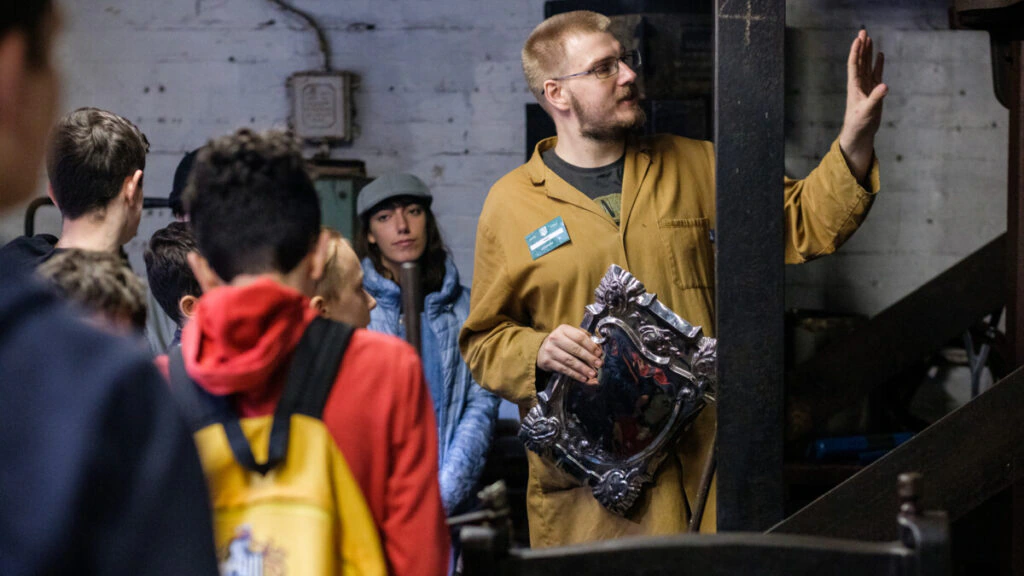 A man in a brown coverall holds an ornate metal frame, speaking to a group of people in a workshop. Some attendees listen attentively, and machinery is visible in the background.