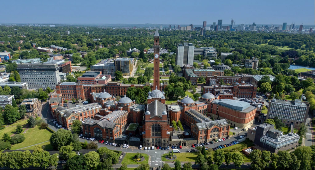 Aerial view of a large university campus with a central clock tower, red brick buildings, green trees, and city skyline in the background.