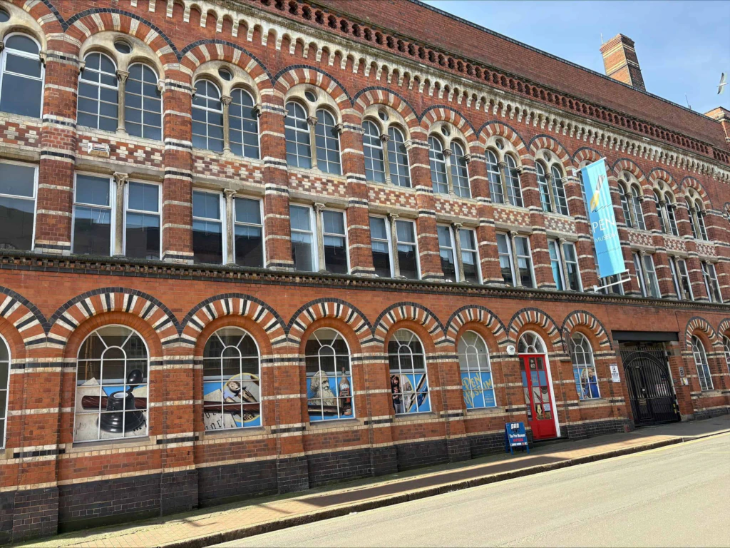 A historic red-brick building with arched windows, decorative brickwork, a blue vertical banner, and a red door. Street and sidewalk are visible in front.