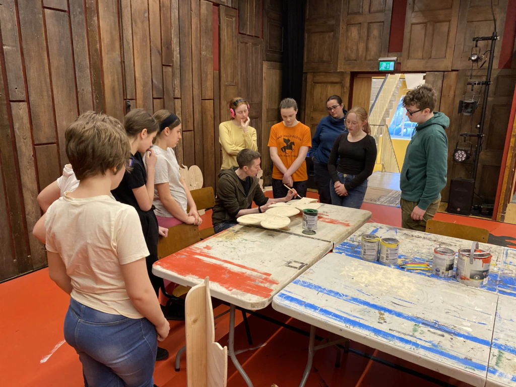 A group of people stand around a table covered in paint cans and supplies, watching someone seated work on a wooden project in a room with wooden walls.