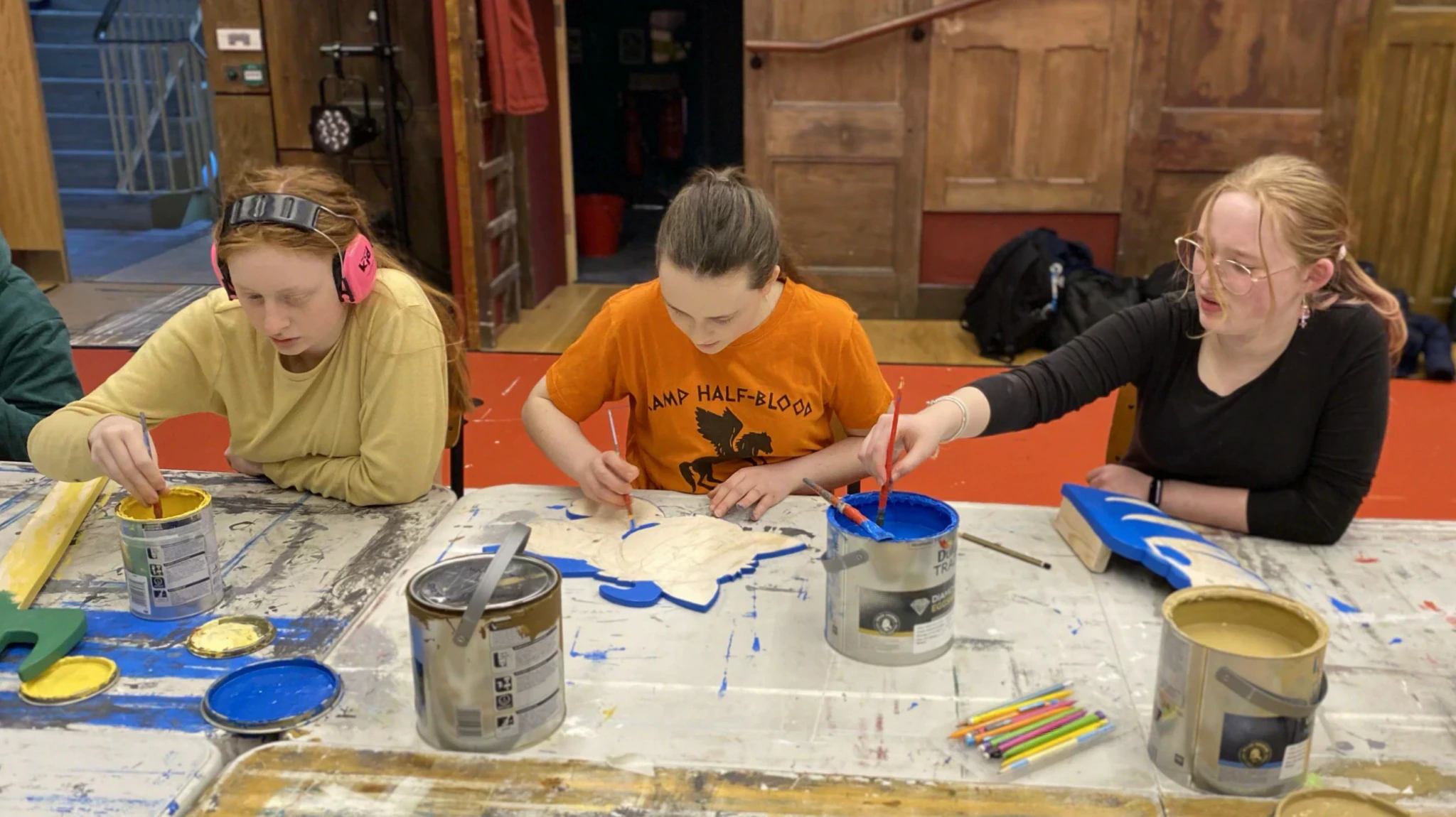 Four young people sit at a table painting wooden props with blue, gold, and silver paints. Art supplies, paint cans, and brushes are scattered across the table in a creative workshop setting.