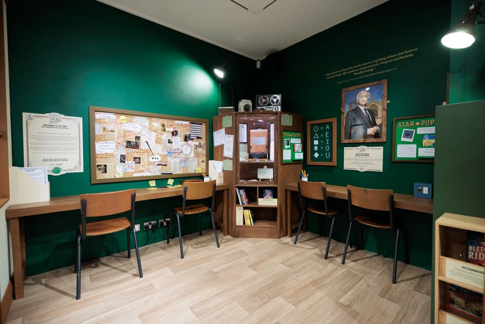 A cozy study room with green walls, a bulletin board, bookshelves, three chairs at a long desk, and a portrait above more notes and papers. Warm lighting and wooden floor create a welcoming atmosphere.