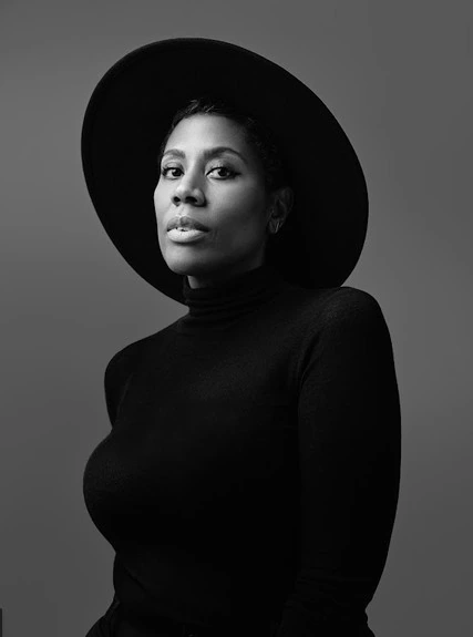 Black and white portrait of a person wearing a wide-brimmed hat and a turtleneck, gazing confidently at the camera against a plain background.