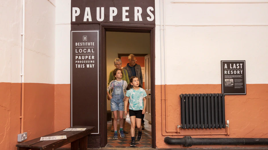 Two children and two adults walk through a doorway labeled “Paupers” in an old building, with signs indicating pauper processing and historical context. The group appears to be visiting a museum exhibit.