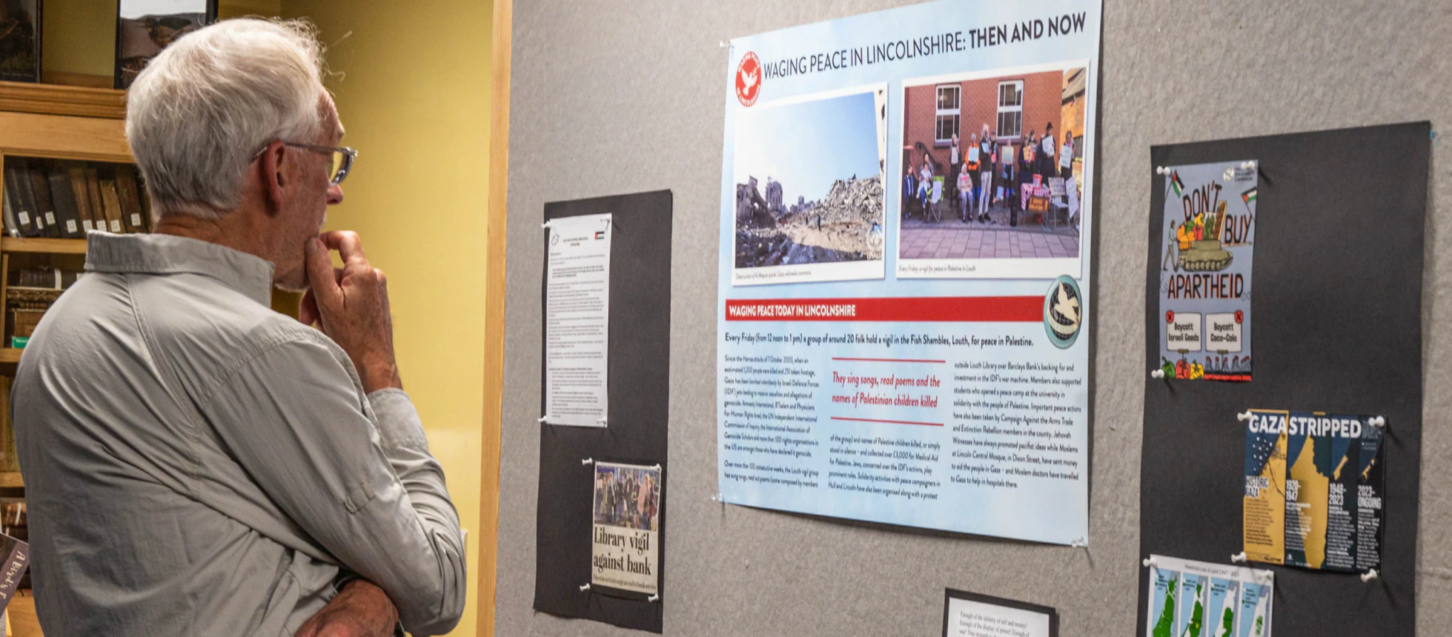 An elderly man stands with his hand on his chin, thoughtfully reading a display board about peace in Lincolnshire. The board has photos, articles, and posters related to activism and historical events.