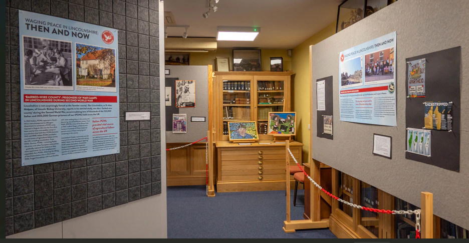 A museum exhibit features historical posters, photos, and documents on display panels, with a wooden display case and bookshelves in the background and a red-and-white chain barrier in the foreground.