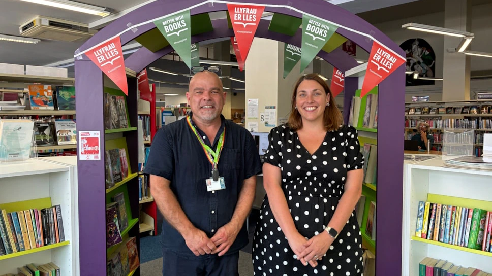 Two adults stand and smile in front of a purple archway decorated with bunting inside a library, surrounded by bookshelves filled with books.