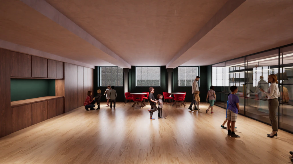 Children and adults interact in a spacious, modern room with wood floors, large windows, and red chairs. Some people stand, others sit or crouch, creating a lively atmosphere in the well-lit area.