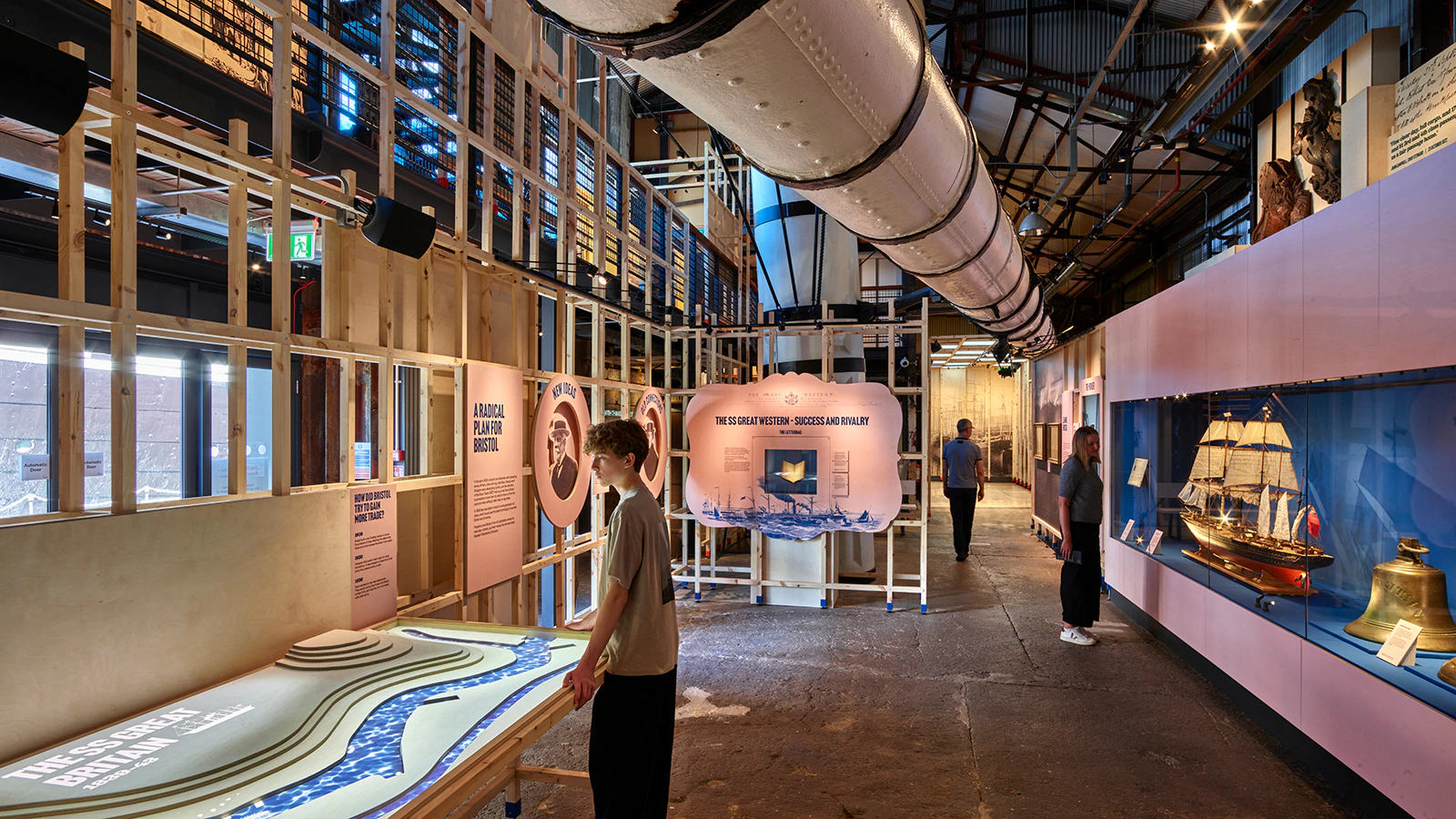 A modern museum exhibit features interactive displays, historical ship models, and visitors exploring. The space has exposed beams, informative panels, and a large overhead pipe running through the center.