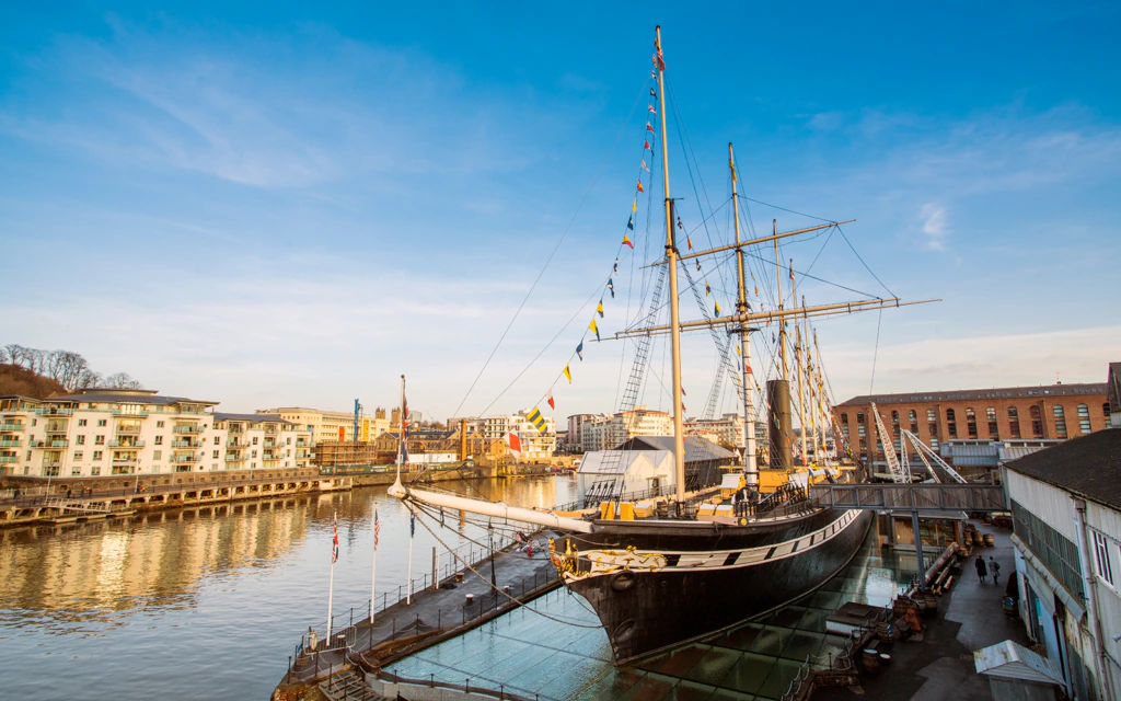 Historic tall ship docked at a waterfront museum, with colorful flags on its masts, surrounded by modern buildings and calm water under a clear blue sky.