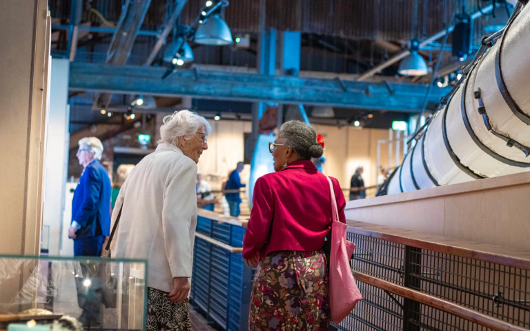 Two elderly women, one in a white jacket and one in a bright pink jacket, stand and talk inside a museum exhibit, with a large industrial machine displayed beside them. Other visitors are visible in the background.