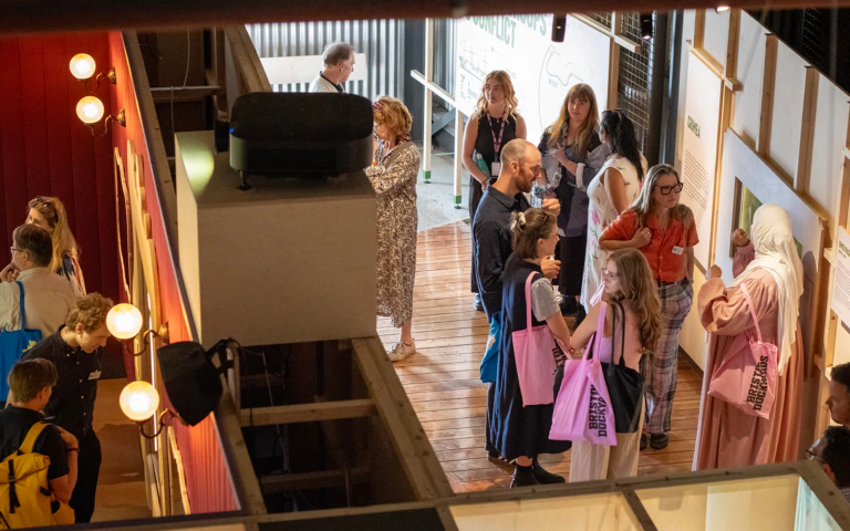 A group of people stand and chat in a brightly lit indoor gallery space, some holding pink tote bags. They appear to be attending an event or exhibition.