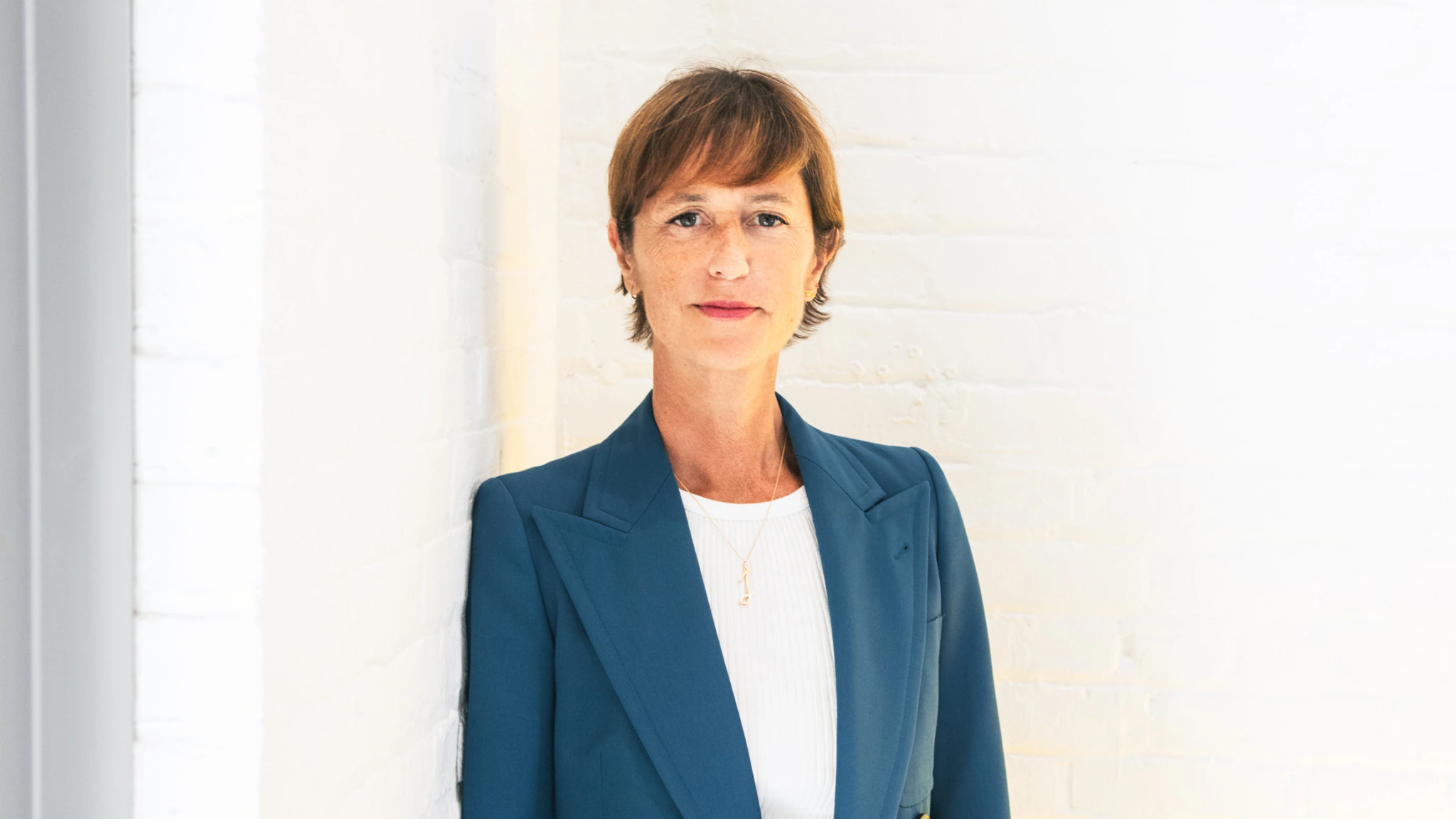 A woman with short brown hair in a teal blazer and white top stands against a white brick wall, looking at the camera with a neutral expression.