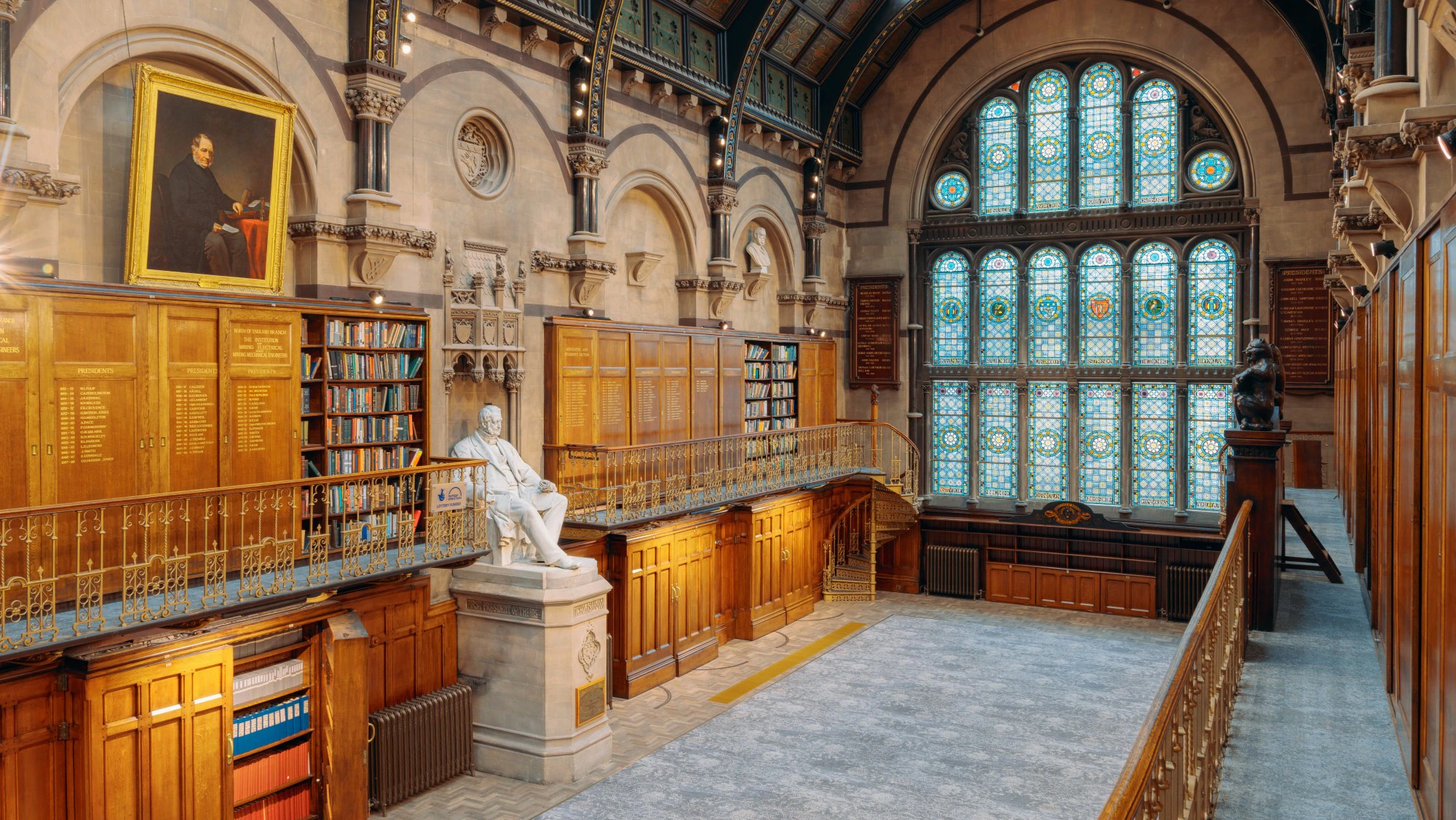 A grand library hall with tall stained glass windows, wooden bookshelves, a statue of a seated figure, and a large portrait on the wall above the bookshelves. The ceiling is arched and ornate.