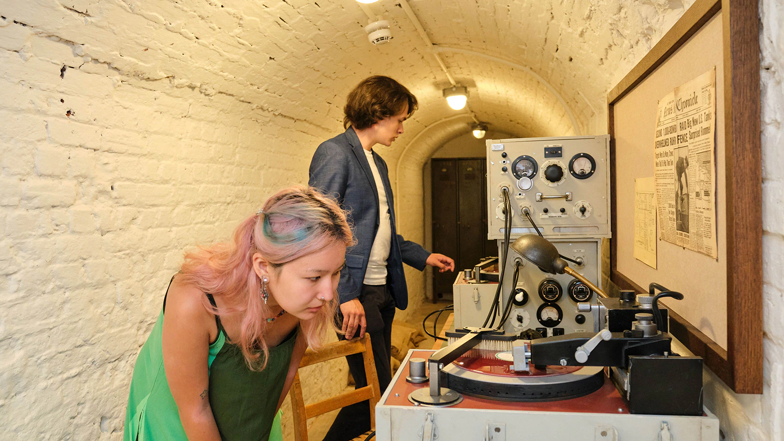 A woman with pink hair leans over vintage audio equipment while a man in a blue jacket stands nearby in a brick-walled room with arched ceiling and framed newspaper.