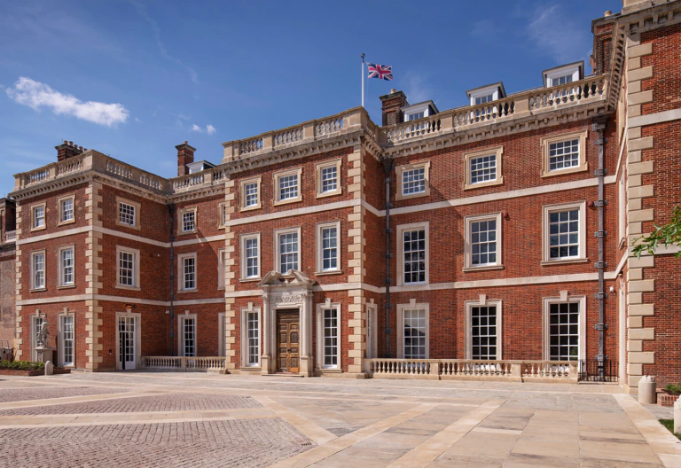 A large, historic brick building with tall windows, stone accents, and a British flag flying on top. The entrance features columns and steps. The sky is clear and blue.
