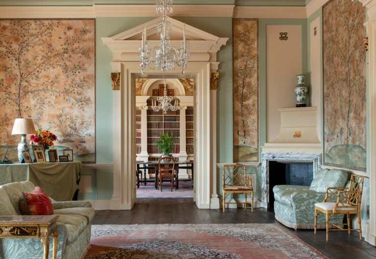 Elegant living room with floral wallpaper, chandeliers, classic fireplace, green sofas, gold accents, and a view into a formal dining room with bookshelves and a central plant. Soft natural light fills the space.