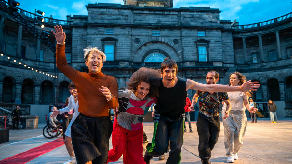 A group of energetic performers runs toward the camera on an outdoor stage, with a historic building and blue sky in the background.