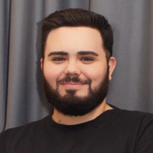 A person with dark hair and a beard smiles while wearing a black Dickies shirt. They are standing in front of gray curtains.