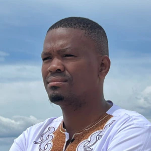 A man in a white patterned shirt stands on a beach with his arms crossed, looking thoughtful. The sea, boats, and buildings are visible in the background under a partly cloudy sky.