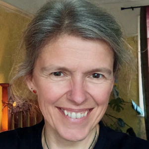 A smiling woman with light hair pulled back, wearing a black shirt and a necklace, stands indoors with greenish walls and a window with maroon curtains in the background.