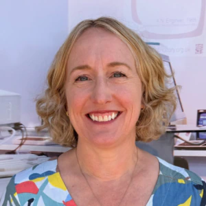 A woman with curly blonde hair, wearing a colorful patterned shirt and a necklace, smiles at the camera. The background shows computer equipment and a blurred office setting.